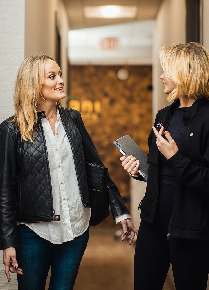 Two women conversing in a modern office hallway.